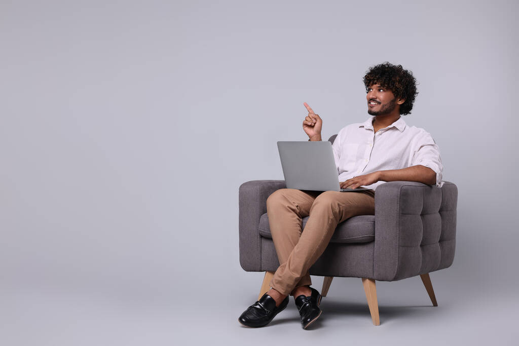Hombre sonriente con portátil sentado en sillón sobre fondo gris claro, espacio para el texto - Foto, imagen