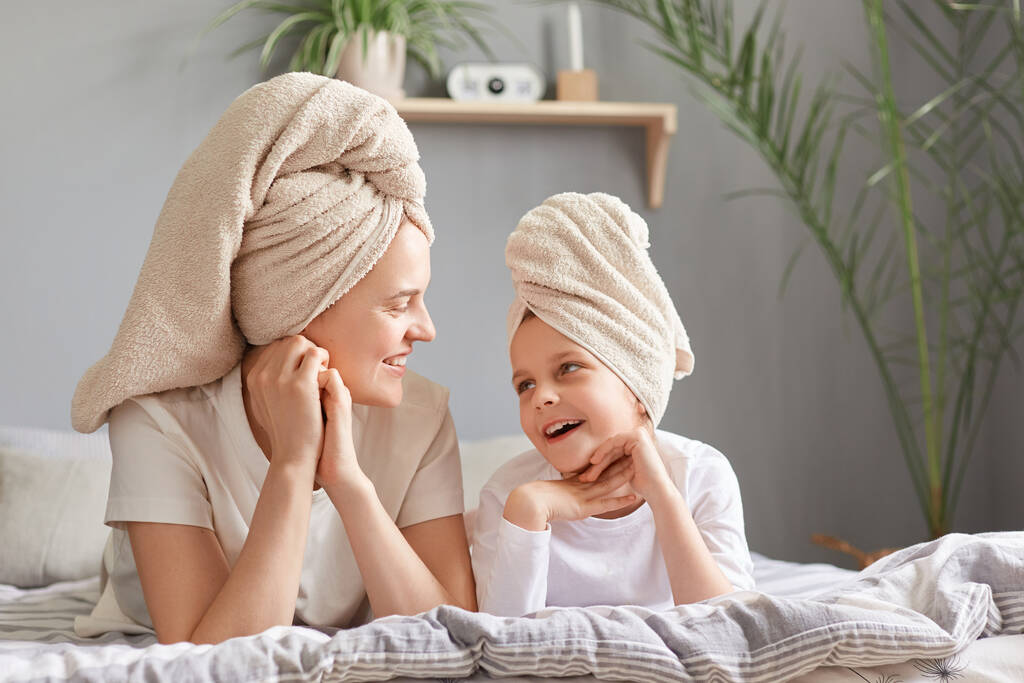 Πορτρέτο της ελκυστικής γυναίκας με το παιδί της μετά τη λήψη δείχνει με το παιδί της, μητέρα και κόρη ξαπλωμένη στο κρεβάτι φορώντας πετσέτα ποζάροντας στο υπνοδωμάτιο στο σπίτι. - Φωτογραφία, εικόνα