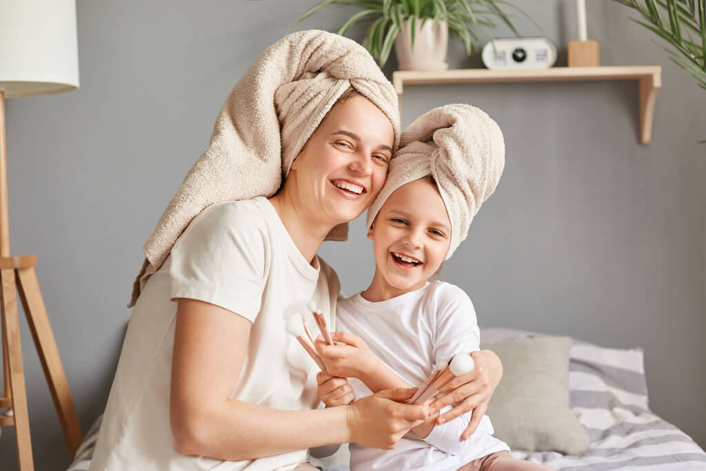 Χαρούμενη γυναίκα αγκαλιάζει το μικρό της παιδί μητέρα και κόρη σε πετσέτες ποζάροντας στην κρεβατοκάμαρα κοιτάζοντας την κάμερα εκφράζοντας τις διαδικασίες ευτυχίας ομορφιάς μαζί. - Φωτογραφία, εικόνα