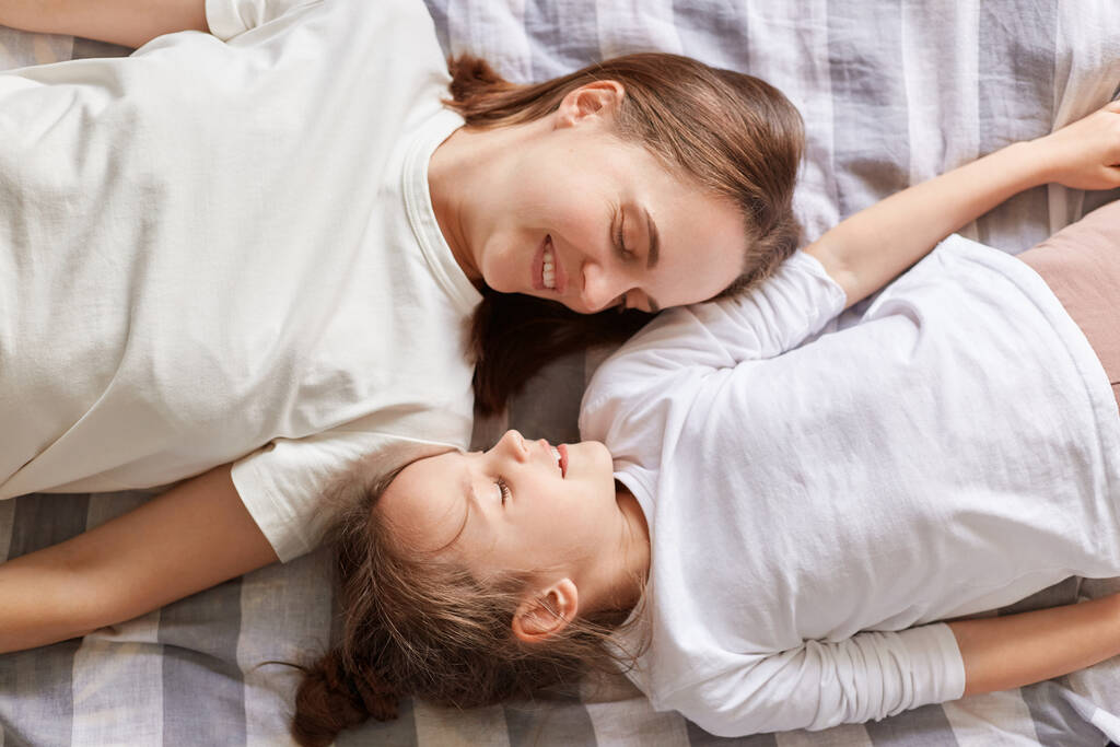 Sorrindo encantadora mãe e filha descansando na cama desfrutando de fim de semana falando de manhã vestindo camisetas brancas feliz relação familiar maternidade. - Foto, Imagem