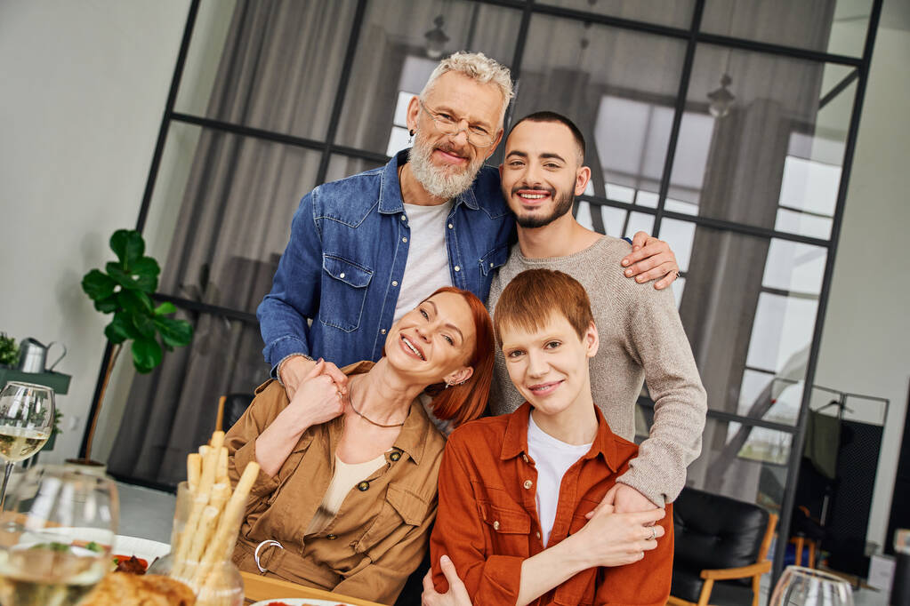 fröhlicher schwuler Mann mit Eltern und Freund, die sich umarmen und in die Kamera lächeln, in der Nähe des servierten Abendessens im Wohnzimmer - Foto, Bild