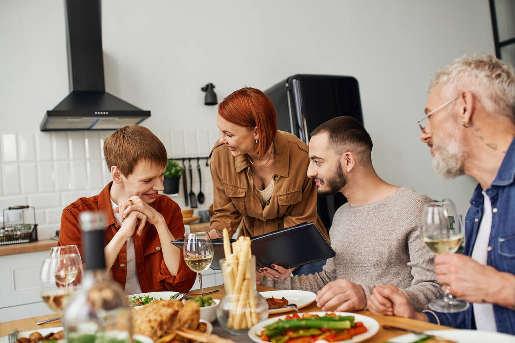 gaie rousse femme montrant album photo de famille à sourire fils avec gay copain près délicieux souper servi dans la cuisine - Photo, image
