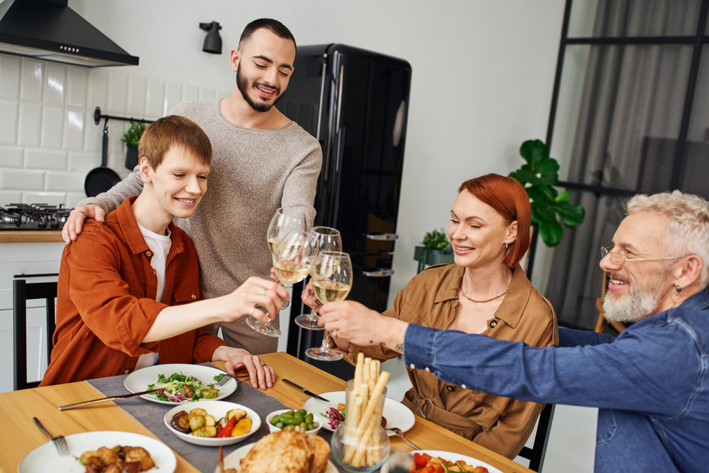 gay feliz homem clinking vinho copos com namorado e pais perto delicioso ceia no cozinha - Foto, Imagem