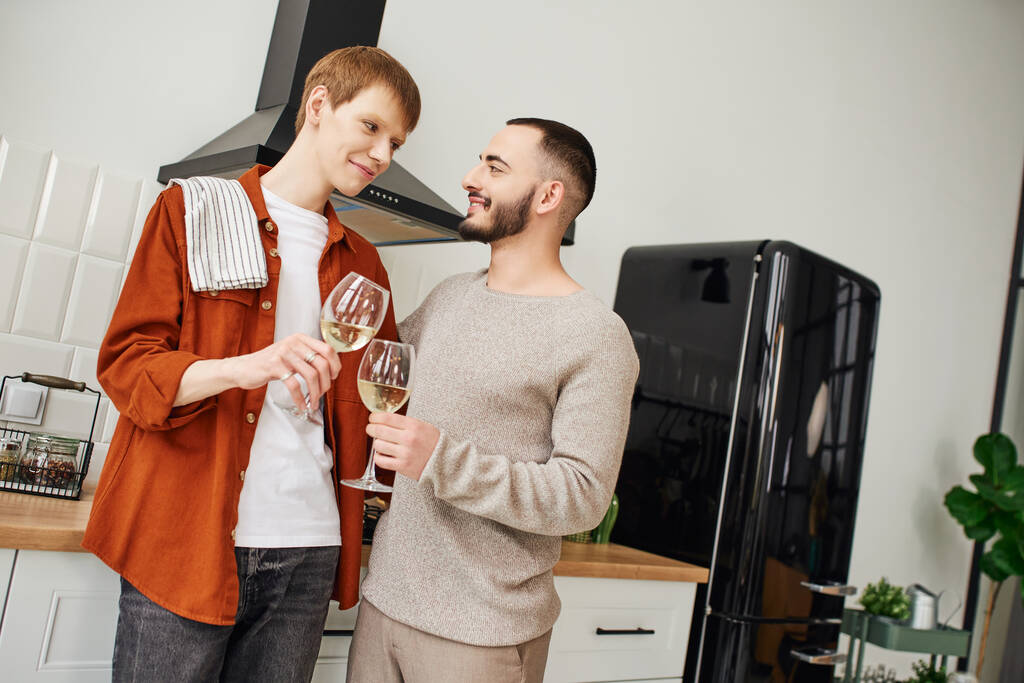 happy gay couple clinking wine glasses and looking at each other in kitchen - Photo, Image
