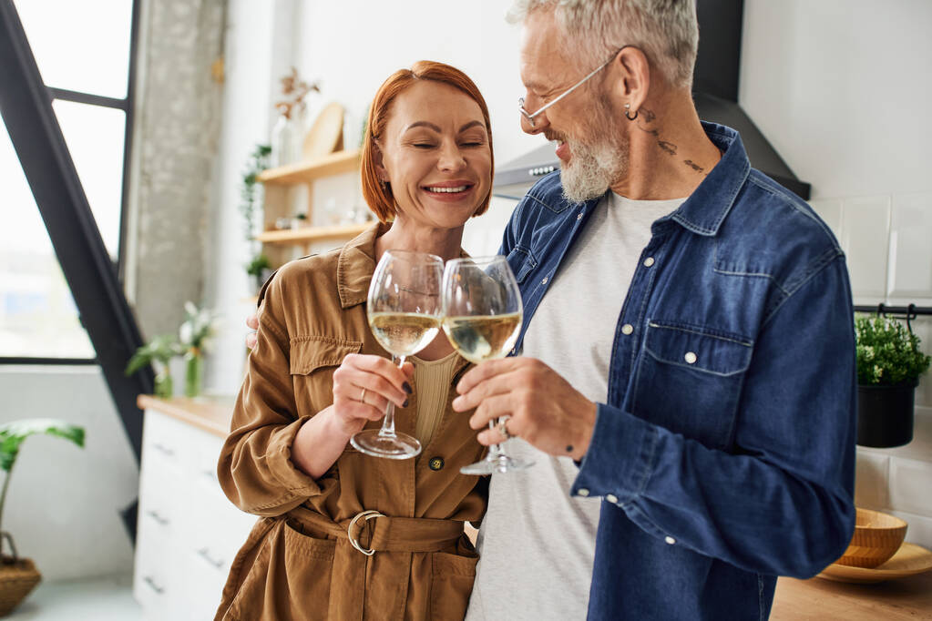 veselý a stylový pár středního věku cinkání sklenice na víno v kuchyni - Fotografie, Obrázek