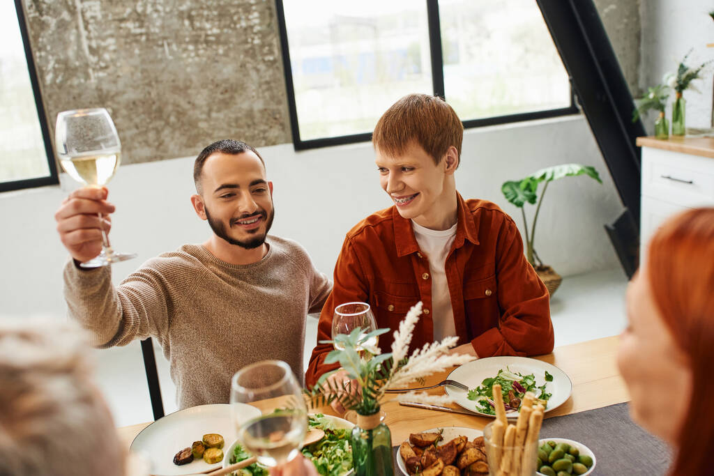 barbudo hombre tostando con vino cerca feliz gay pareja y delicioso familia cena en moderno cocina - Foto, Imagen