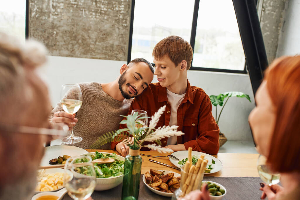 zadowolony gej człowiek z wino szklanka opierając się na ruda chłopak w pobliżu niewyraźne rodzice i pyszne kolacja w kuchnia - Zdjęcie, obraz