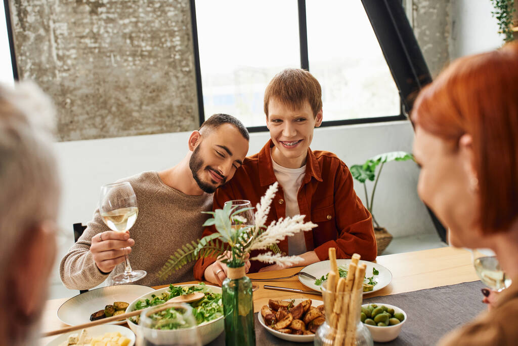 Bir bardak şarapla neşeli sakallı bir adam mutfaktaki aile yemeğinde mutlu eşcinsel eşine yaslanıyor. - Fotoğraf, Görsel