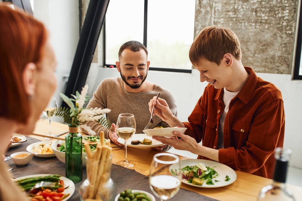 веселый рыжий мужчина, подающий еду рядом с гей-партнером и размытая мать во время семейного ужина на кухне - Фото, изображение