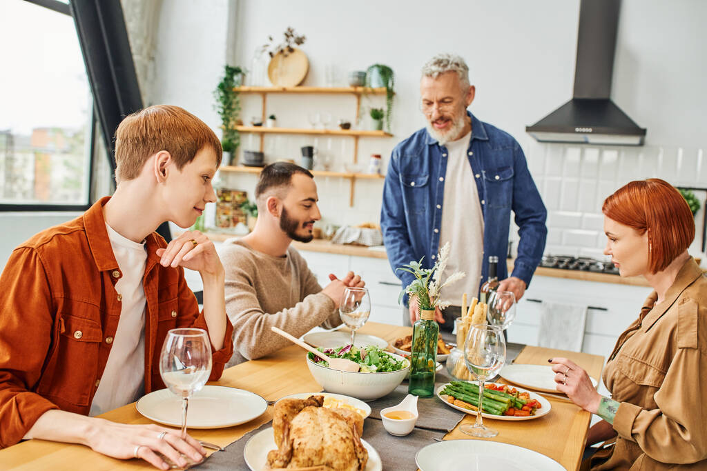 man met baard staan met fles wijn in de buurt van zoon met gay partner tijdens familie diner in keuken - Foto, afbeelding