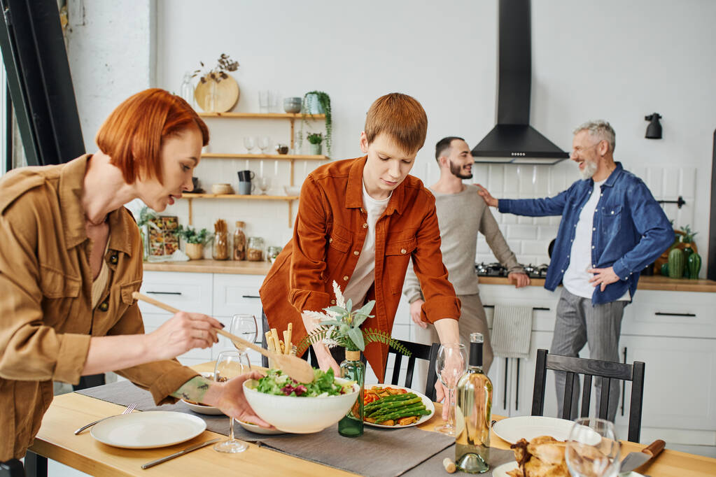 Рыжая женщина смешивает салат рядом с сыном накрывая семейный ужин на столе на кухне - Фото, изображение
