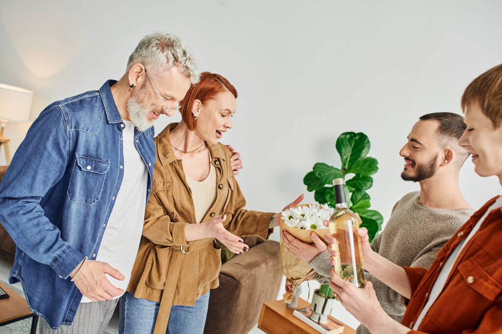 junges schwules Paar besucht glückliche Eltern zu Hause, während es Blumen und eine Flasche Wein in der Hand hält - Foto, Bild