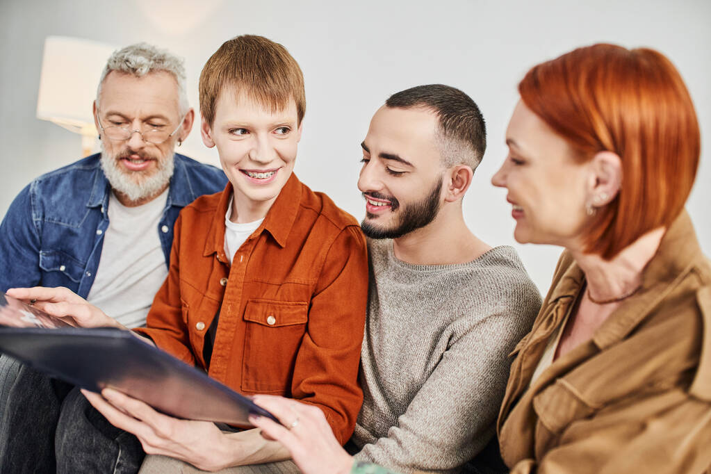 радісний гей чоловік посміхається біля хлопця і батьків, тримаючи фотоальбом у вітальні
 - Фото, зображення