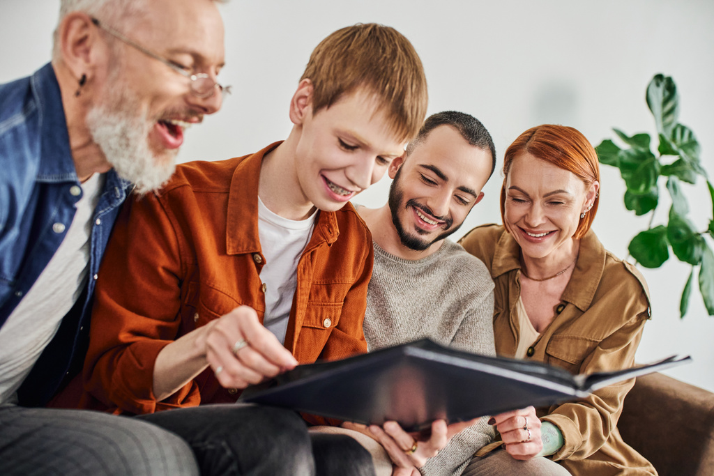 young gay man looking at photo album near bearded boyfriend and cheerful parents - Photo, Image