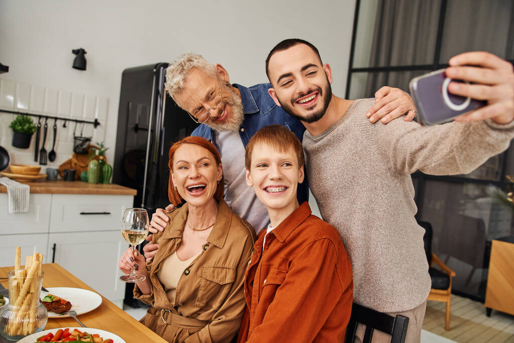 Χαρούμενος γκέι άντρας που βγάζει selfie με το αγόρι του και χαρούμενους γονείς κατά τη διάρκεια του οικογενειακού δείπνου στο σπίτι - Φωτογραφία, εικόνα