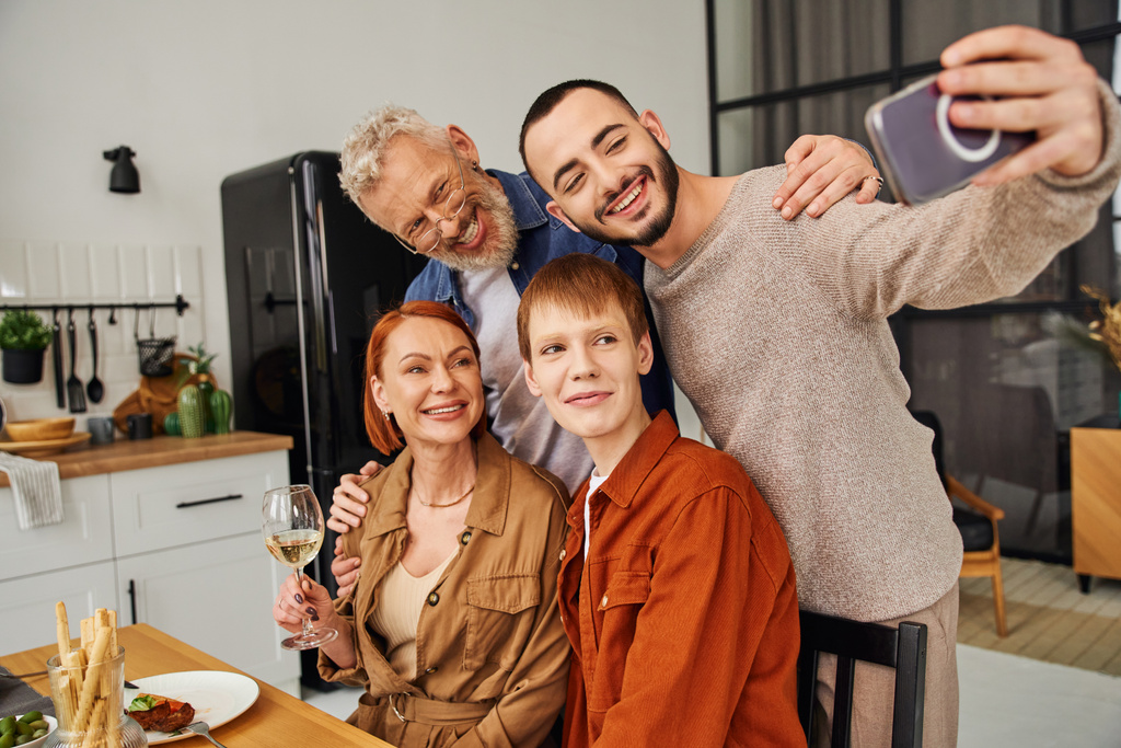 веселый мужчина делает селфи с гей-партнером и родителями во время семейного ужина на кухне - Фото, изображение