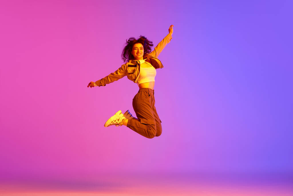 Mladá dívka, hip-hop tanečník ve sportovním stylu oblečení skákání proti gradient růžové fialové pozadí v neonovém světle. Rozkošné. Koncept současného tance, mládí, hobby, akce a pohybu - Fotografie, Obrázek