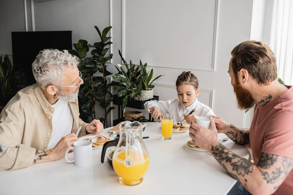 Tätowierte schwule Eltern schauen auf preteen Tochter mit Frühstück zu Hause  - Foto, Bild