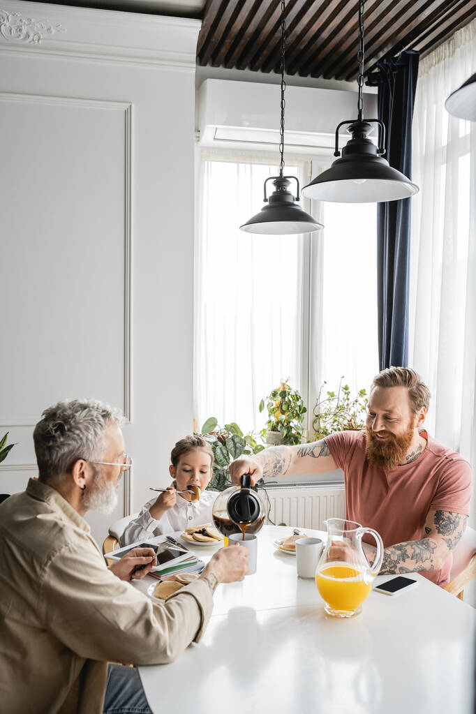 Uśmiechnięty mężczyzna nalewający kawę w pobliżu córki i partnera geja podczas śniadania w domu  - Zdjęcie, obraz