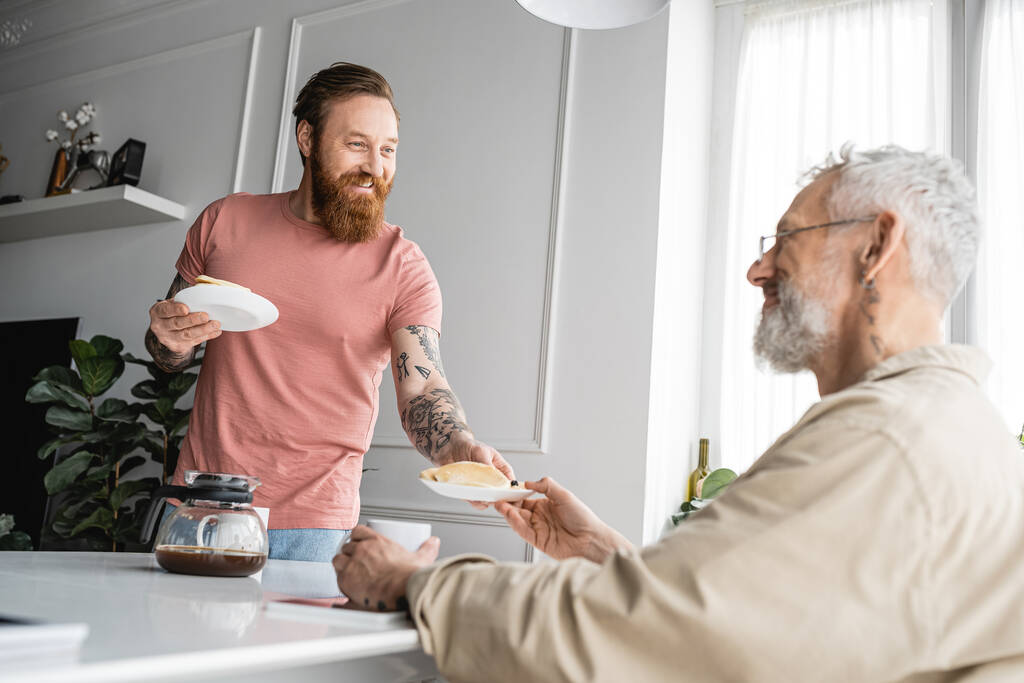 Tätowierter schwuler Mann lächelt, während er seinem Partner zu Hause Pfannkuchen gibt  - Foto, Bild