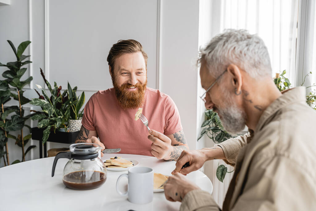 Hombre homosexual tatuado mirando a su pareja durante el desayuno en casa  - Foto, Imagen