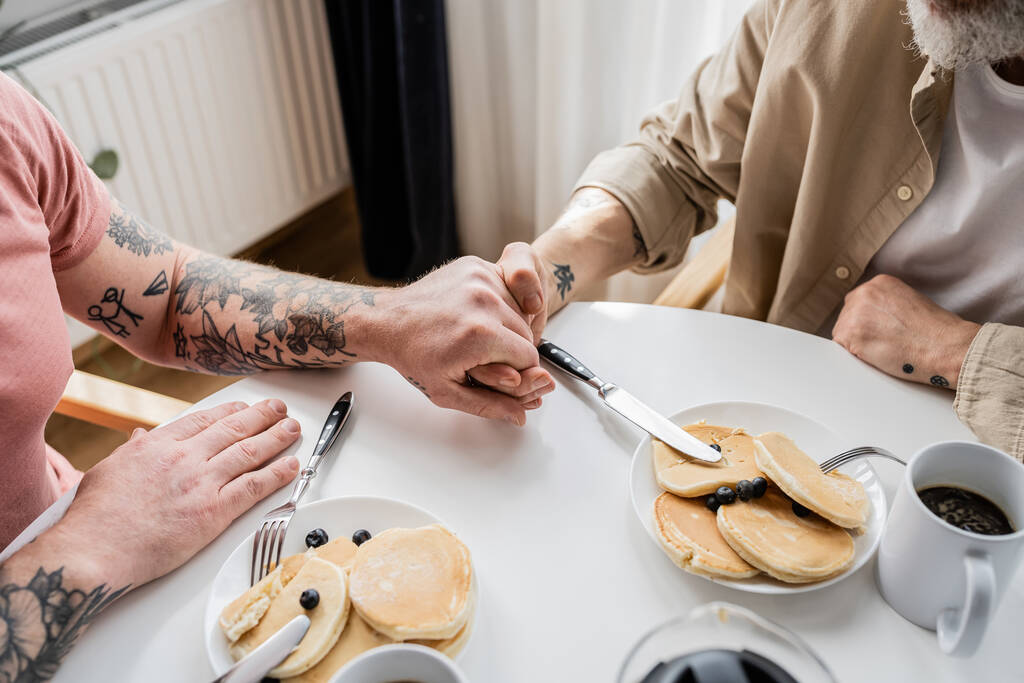 Gewassen uitzicht van getatoeëerd gay paar houden handen in de buurt van pannenkoeken en koffie thuis  - Foto, afbeelding