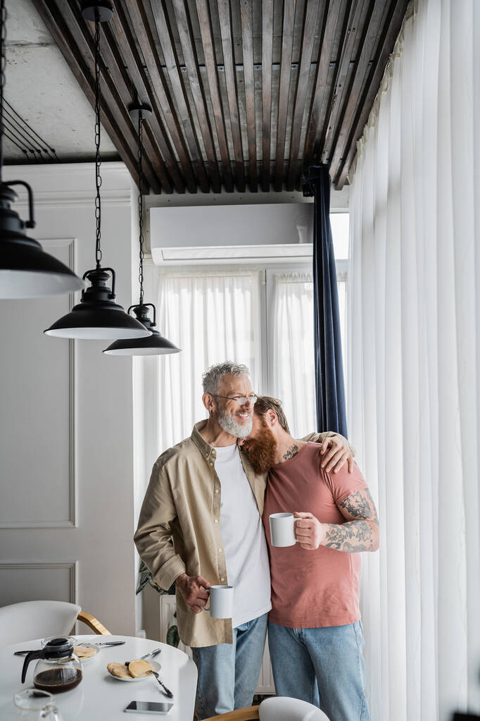 Беззаботный гомосексуальный мужчина обнимает татуированного партнера с кофе возле завтрака дома  - Фото, изображение