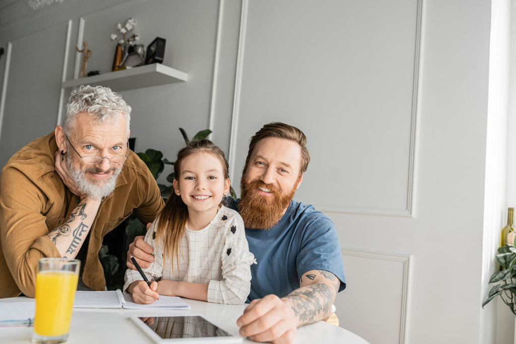 Smiling gay parents with daughter looking at camera while doing homework at home  - Photo, Image