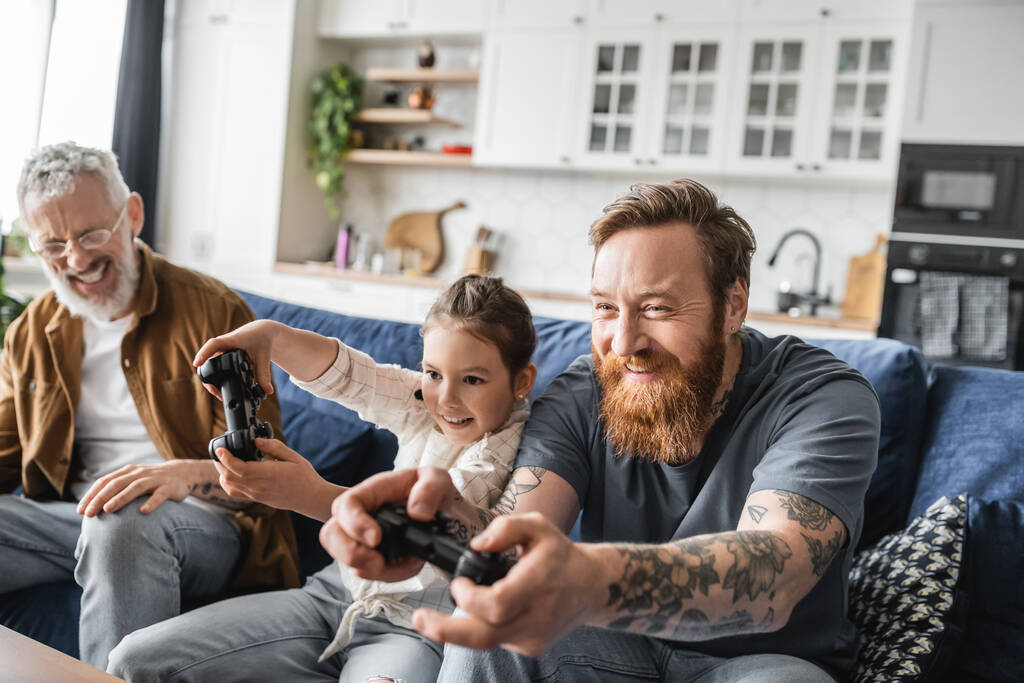 KYIV, UCRANIA - 19 de abril de 2023: Papá homosexual tatuado jugando videojuegos con una hija preadolescente cerca de su pareja en casa  - Foto, imagen