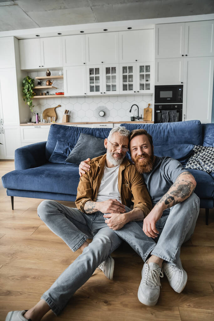 Positiivinen homoseksuaalinen mies halaamassa tatuoitua kumppania lattialla kotona  - Valokuva, kuva