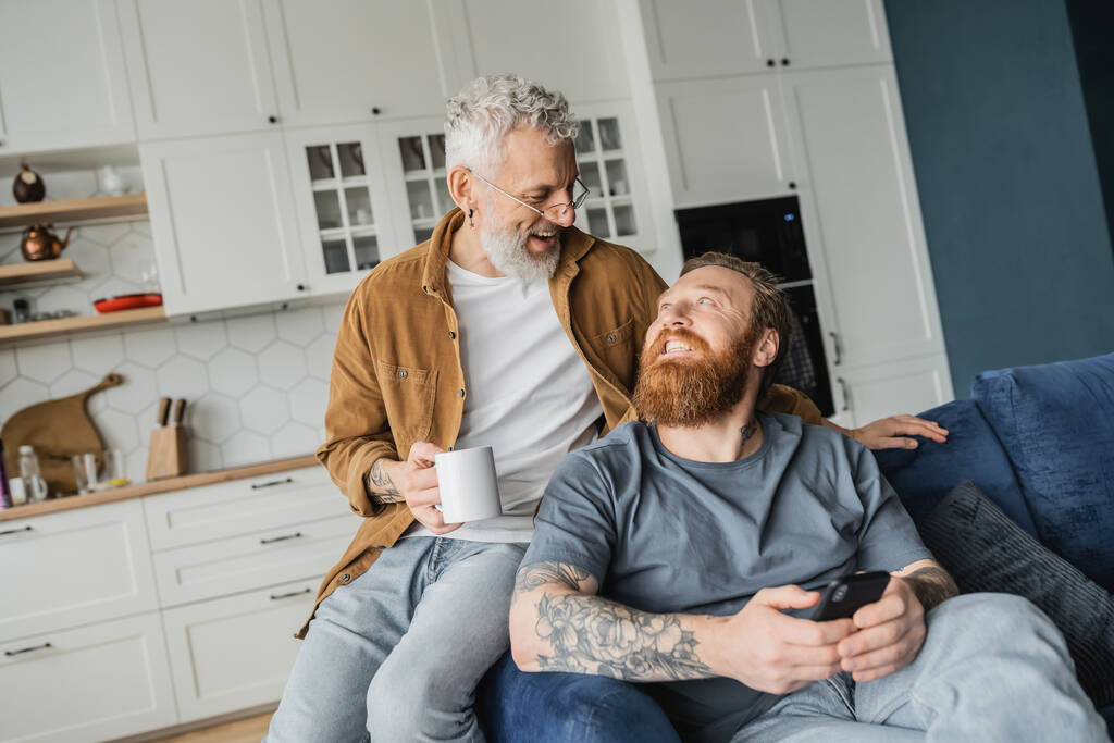 Hymyilevä homo mies, jolla on kahvikuppi lähellä kumppania älypuhelimen kanssa sohvalla kotona  - Valokuva, kuva