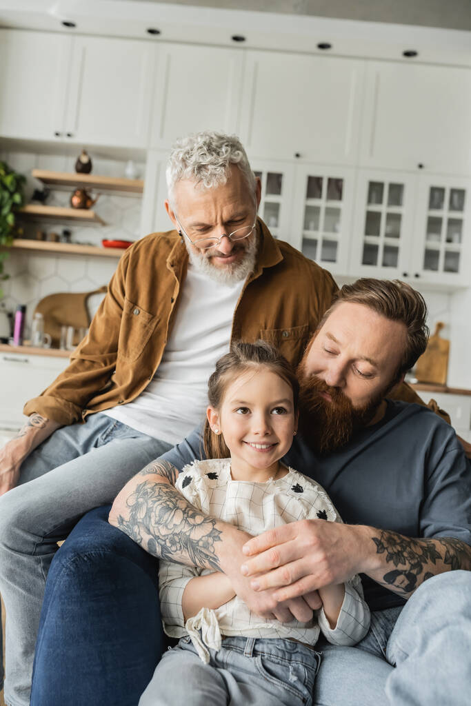 Homosexual parents hugging and looking at smiling daughter at home  - Photo, Image