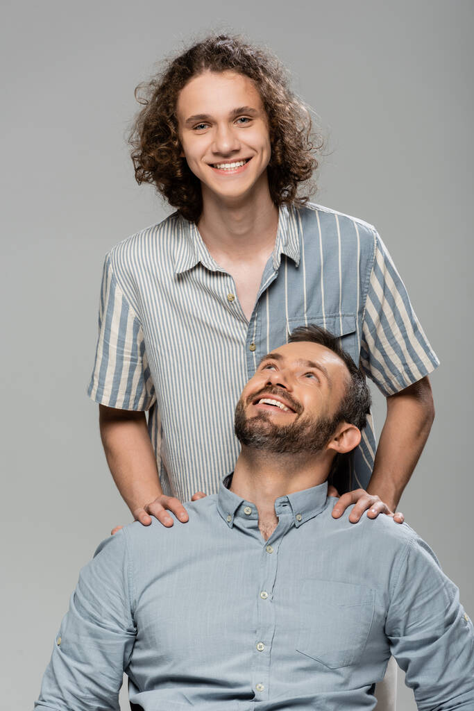 positive father smiling and looking at happy teenage son isolated on grey  - Photo, Image
