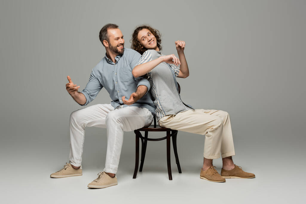 smiling father and teenage son having fun and pushing each other while sitting on same chair on grey  - Photo, Image