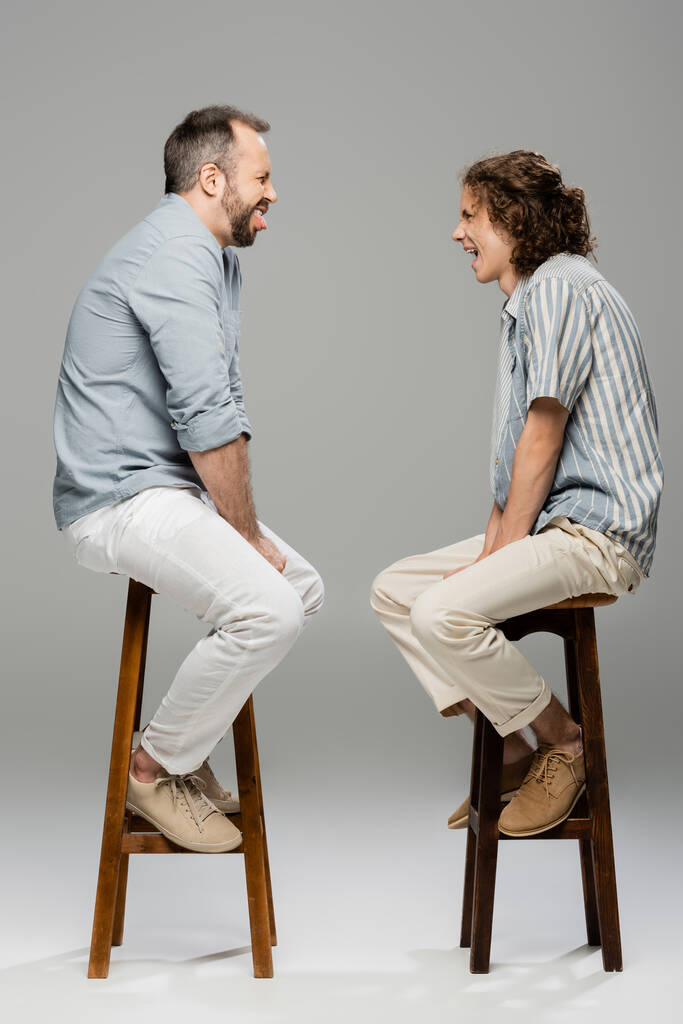 Seitenansicht von lustigen Vater und Teenager-Sohn Grimassen aufeinander, während sie auf Hochstühlen auf grau sitzen  - Foto, Bild