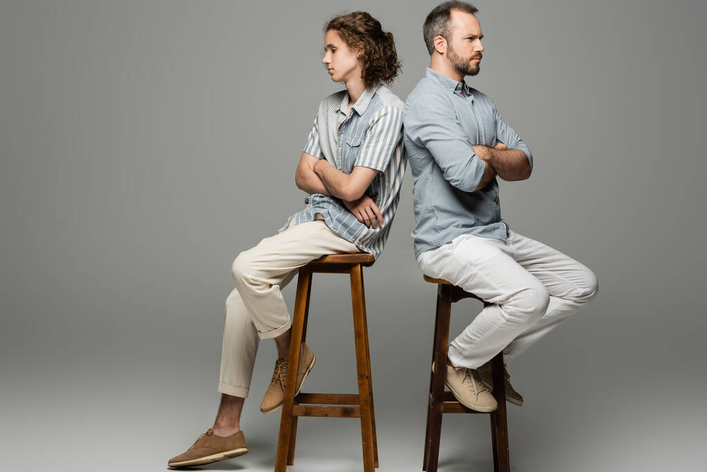 beleidigter Vater und Teenager-Sohn sitzen mit verschränkten Armen Rücken an Rücken auf grau  - Foto, Bild