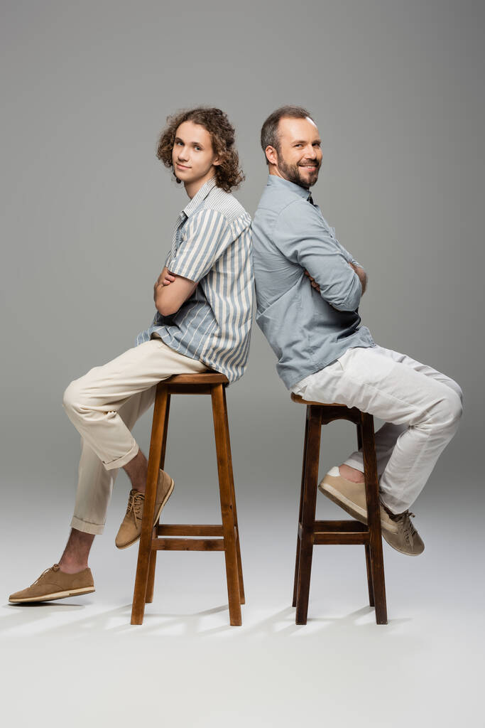 cheerful dad and teenage son sitting with folded arms back to back on grey  - Photo, Image