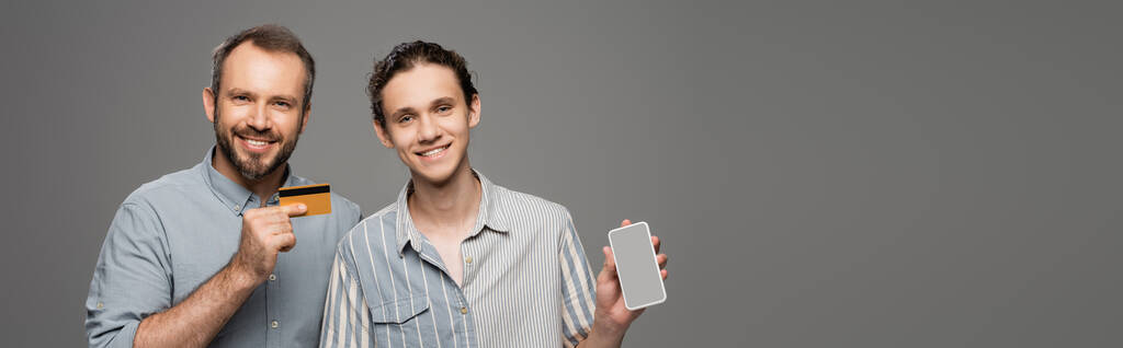 szczęśliwy ojciec trzyma kartę kredytową obok nastoletniego syna ze smartfonem w ręku odizolowanym na szarym, bannerze  - Zdjęcie, obraz