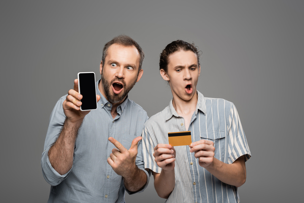 astonished father holding smartphone with blank screen next to shocked teenage son with credit card in hand isolated on grey  - Photo, Image