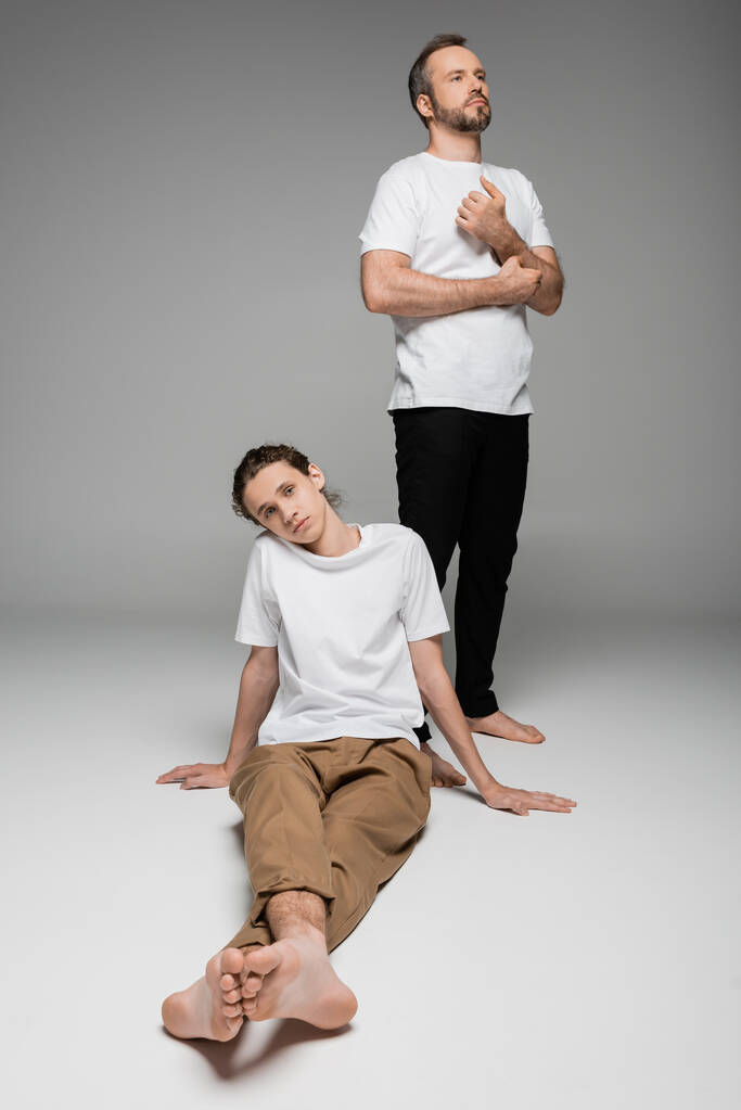longitud completa del padre de pie junto a su hijo adolescente en camiseta blanca en gris  - Foto, Imagen