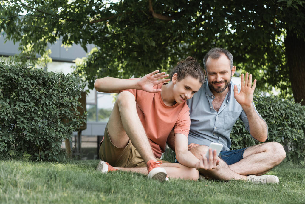 feliz padre e hijo adolescente saludando las manos durante la videollamada mientras están sentados juntos en el parque verde  - Foto, Imagen
