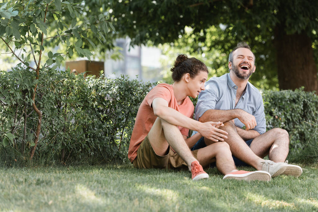 feliz padre riendo mientras está sentado cerca de hijo adolescente en el césped verde en el parque  - Foto, imagen