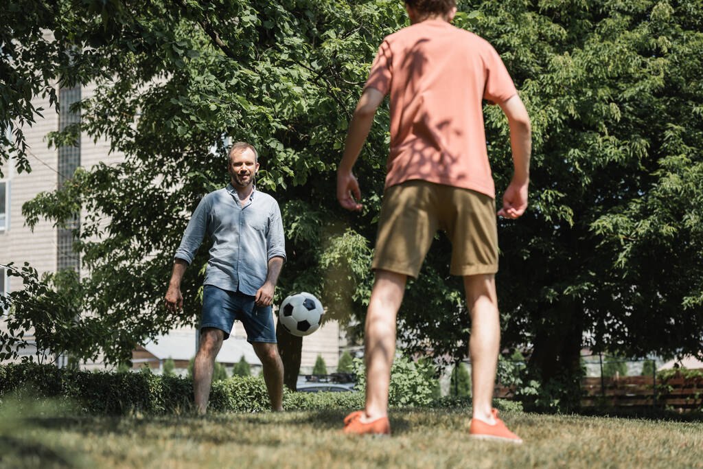 γενειοφόρος άντρας που παίζει ποδόσφαιρο με τον έφηβο γιο του στο πράσινο καλοκαιρινό πάρκο  - Φωτογραφία, εικόνα