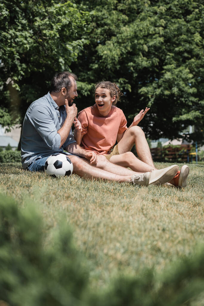 изумленный подросток, сидящий рядом с футбольным мячом с отцом в зеленом летнем парке  - Фото, изображение