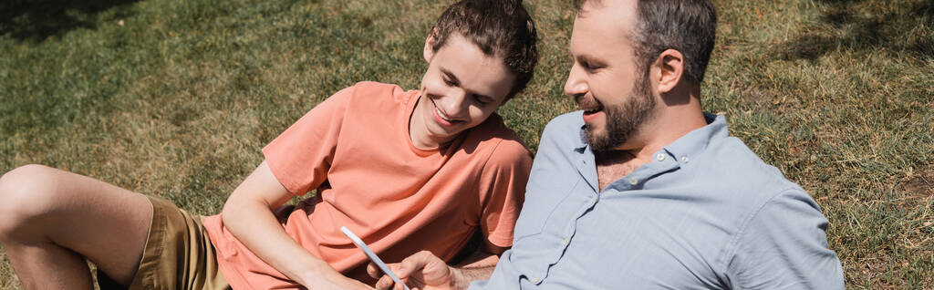 padre barbuto sorridente durante l'utilizzo di smartphone e riposare sul prato vicino figlio adolescente, banner   - Foto, immagini