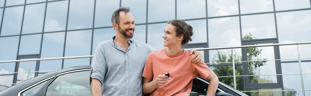 happy bearded man putting hand on shoulder of cheerful teenage son holding car key near automobile, banner  - Photo, Image