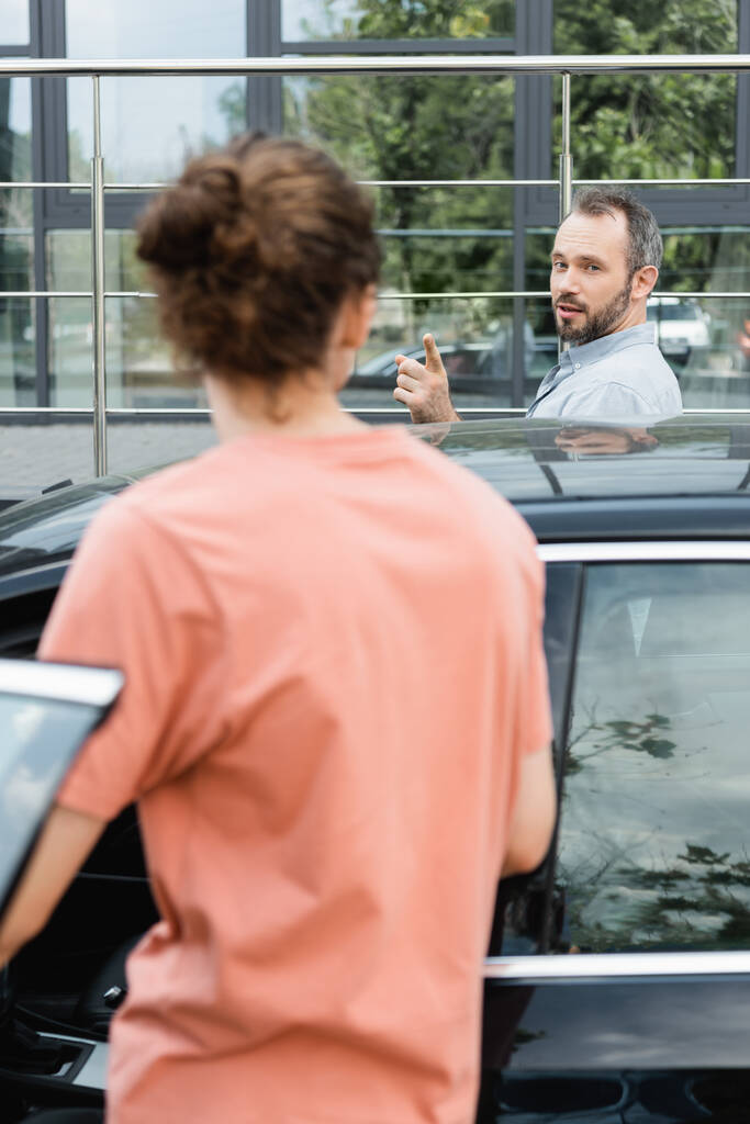 padre barbuto indicando figlio adolescente mentre in piedi insieme vicino auto moderna  - Foto, immagini