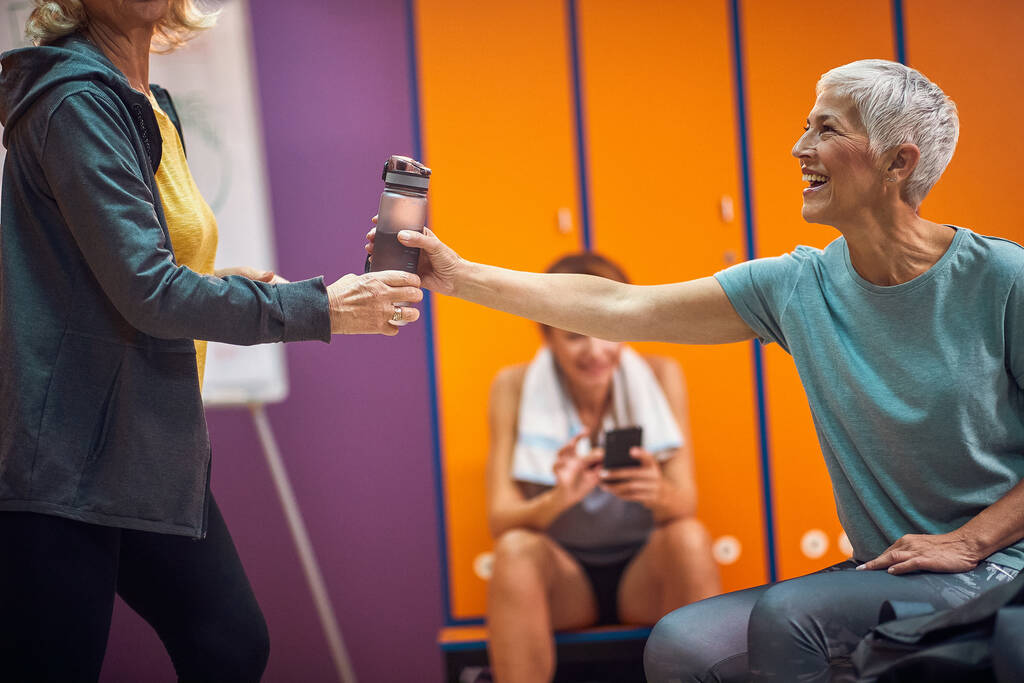 Femme âgée en vêtements de sport offrant à son ami une bouteille d'eau. Groupe de femmes dans les vestiaires de gymnastique ensemble. - Photo, image