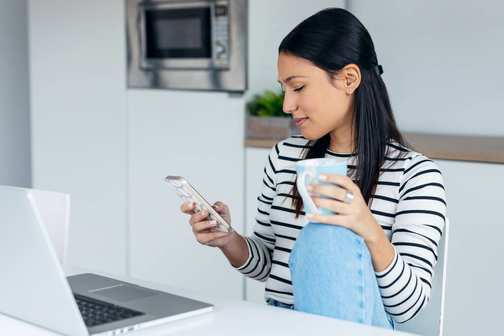Egy gyönyörű, kedves nő, aki üzenetet küld a mobiljával, miközben dolgozik és kávét iszik otthon a konyhában.. - Fotó, kép