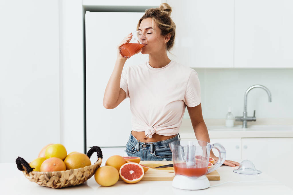 Mujer joven bebiendo zumo de pomelo casero recién exprimido en cocina blanca - Foto, imagen